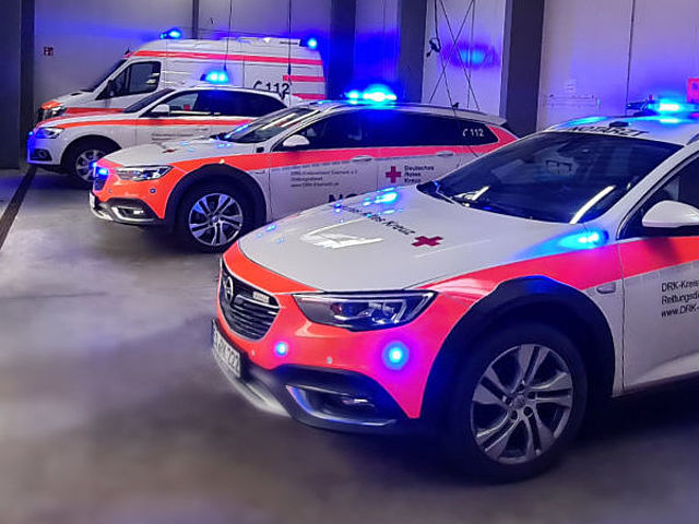 Das Bild zeigt mehrere Rettungs-Fahrzeuge vom Roten Kreuz in einer großen Halle. Vorne stehen zwei moderne, weiße Autos mit roten Streifen. Es sind Notarzt-Einsatz-Fahrzeuge. Ganz hinten steht ein großer, weißer Rettungs-Wagen. Bei allen Fahrzeugen leuchten die blauen Lichter auf dem Dach und an der Vorder-Seite hell. Die Halle ist grau und sauber.