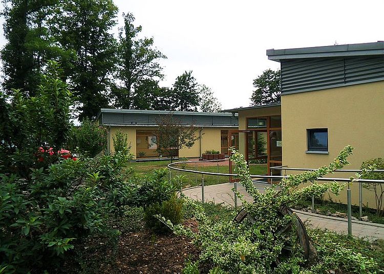 Ein einstöckiges, modernes Gebäude mit hellbeiger Fassade, das von einem gepflegten, halbrunden Garten und grünen Büschen umgeben ist. Ein geschwungener, barrierefreier Weg mit einem Metallgeländer führt zum Eingang. Die Architektur ist offen und hell.